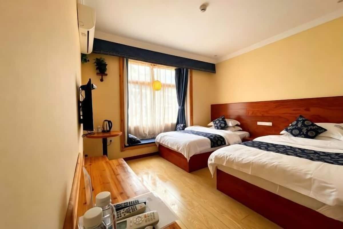 A hotel room with two beds, a wooden floor, a window with curtains, a small table with a kettle, and a wall-mounted air conditioner.