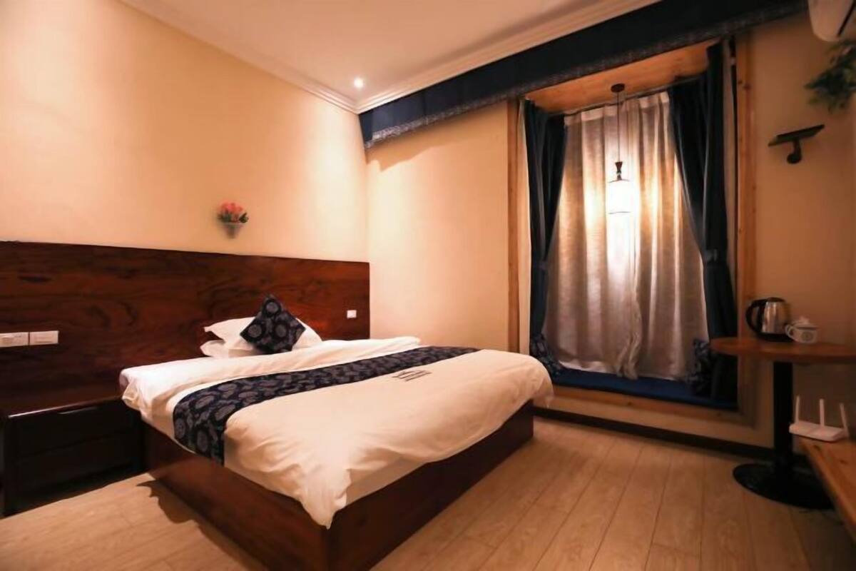 A bedroom with a wooden headboard, a bed with white bedding, a bedside table, a nightstand, a window with curtains, and a small plant on the wall.