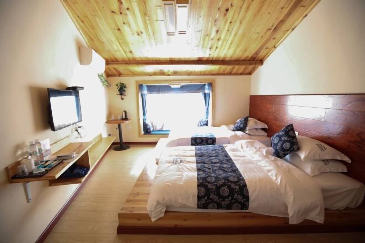 A bedroom with a wooden ceiling, a bed with patterned bedding, a bedside table, a wall-mounted TV, and a window with a view.