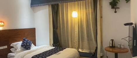 A hotel room with two beds, a small table, a wall-mounted TV, and a window with curtains.