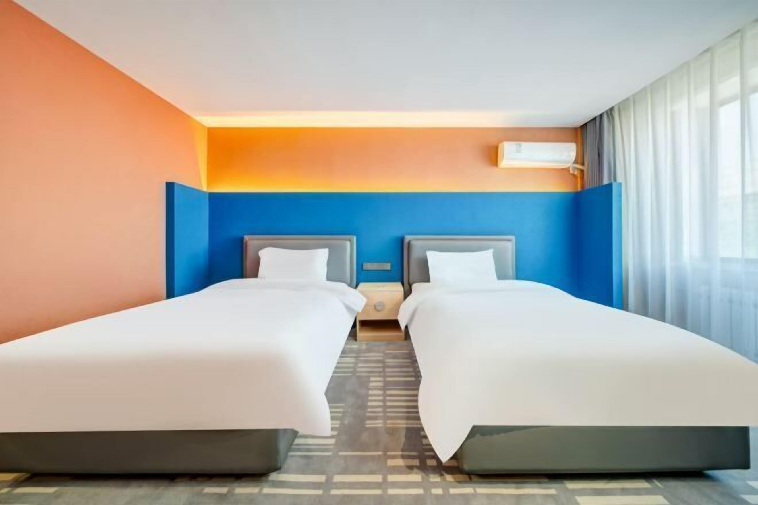 A hotel room with two beds, a nightstand, a wall-mounted air conditioner, and a window with curtains.