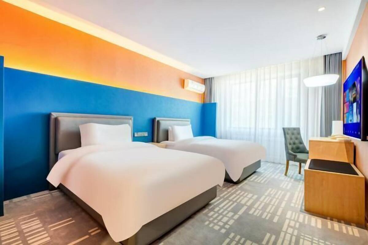A hotel room with two beds, a desk, a chair, a TV, and a window with curtains.