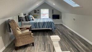 A cozy attic bedroom with a sloped ceiling, a bed with a blue and white patterned bedspread, a wicker chair, a small table with a lamp, and a window with blinds. - Only 2 blocks from all the action on Canal Street! (New Smyrna Beach)