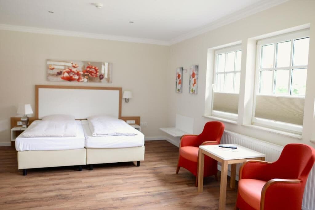 Habitación de hotel con cama doble, una mesita pequeña, dos sillas rojas y una ventana con persianas.