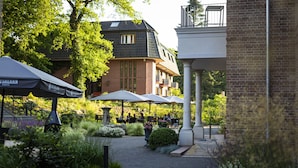 An outdoor café area with tables and umbrellas, surrounded by greenery and a brick building in the background. - Hotel Gut Altona (Dötlingen)