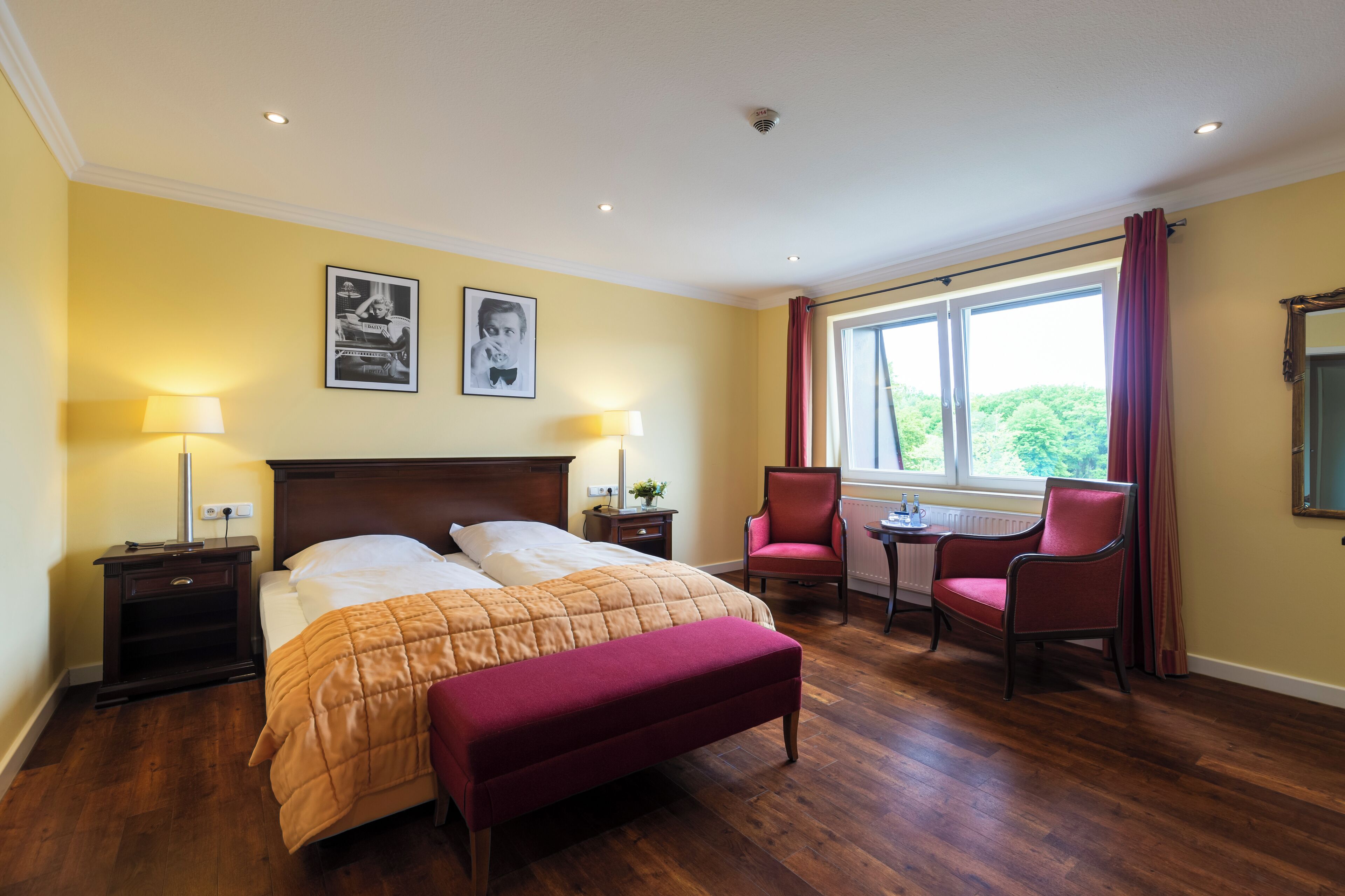 Habitación de hotel con cama, dos sillas, una mesa, un banco, una ventana con cortinas y dos cuadros en la pared.