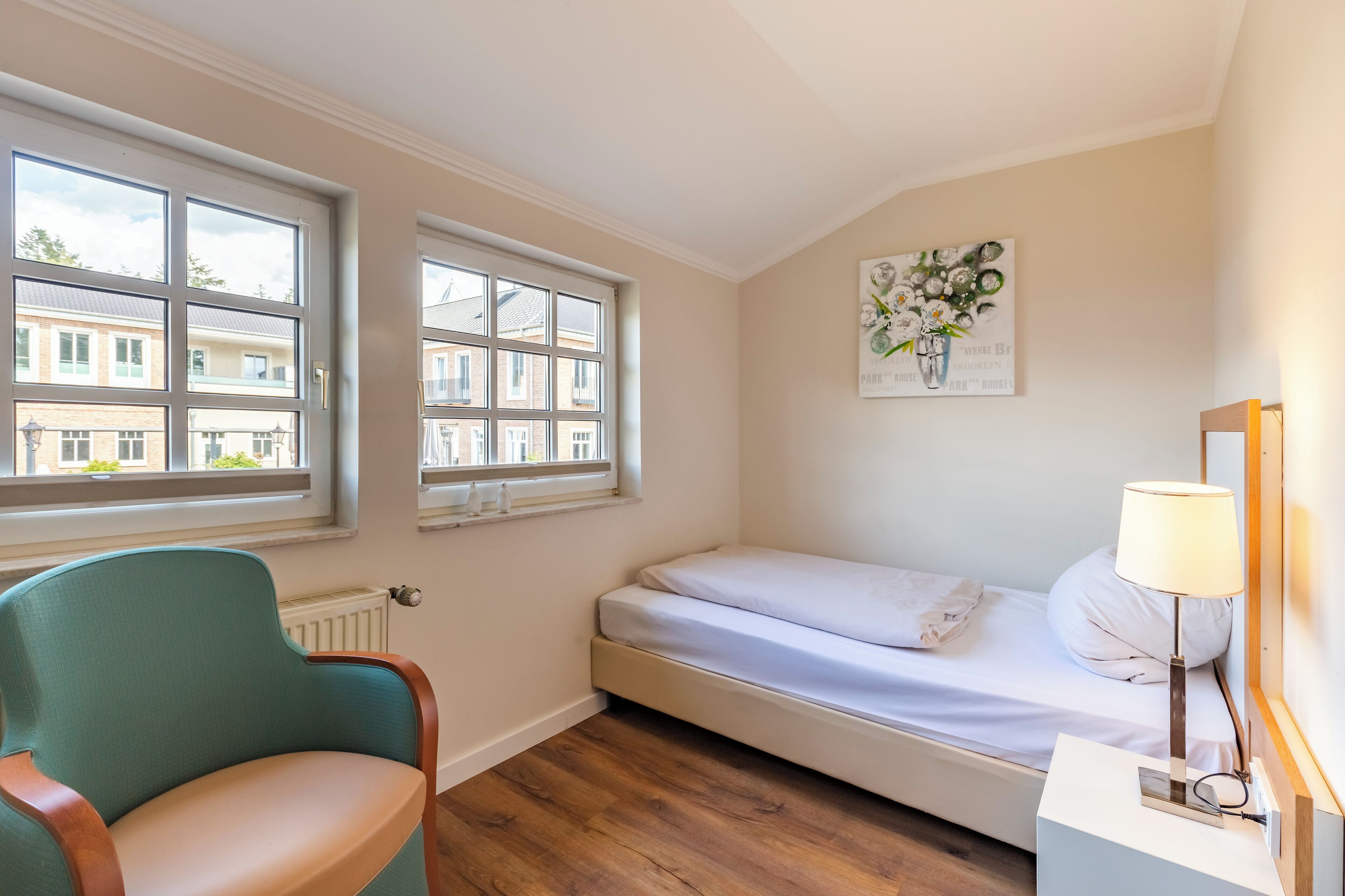 Un dormitorio con una cama individual, una silla, una mesita de noche con lámpara y una ventana con vistas a edificios.