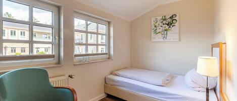 Un dormitorio con una cama individual, una silla, una mesita de noche con lámpara y una ventana con vistas a edificios.