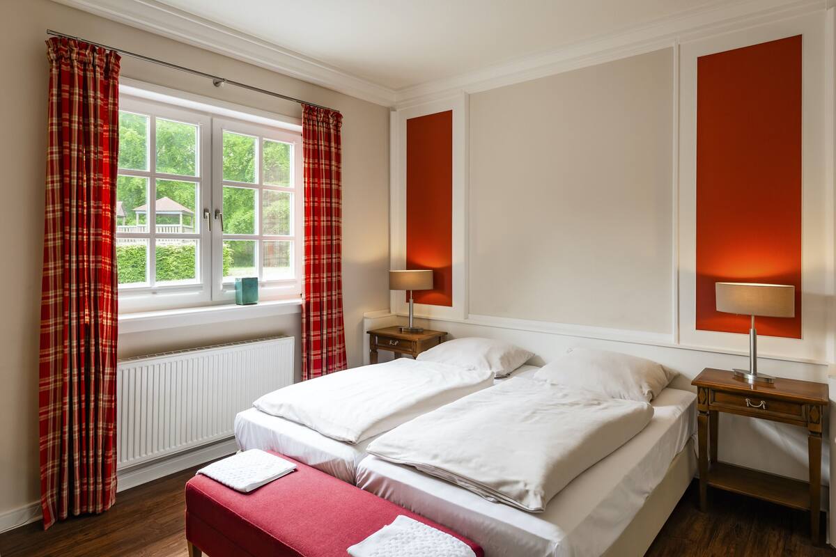 Ein Doppelbett mit weißer Bettwäsche, ein Nachttisch mit Lampe und ein Fenster mit roten Vorhängen.