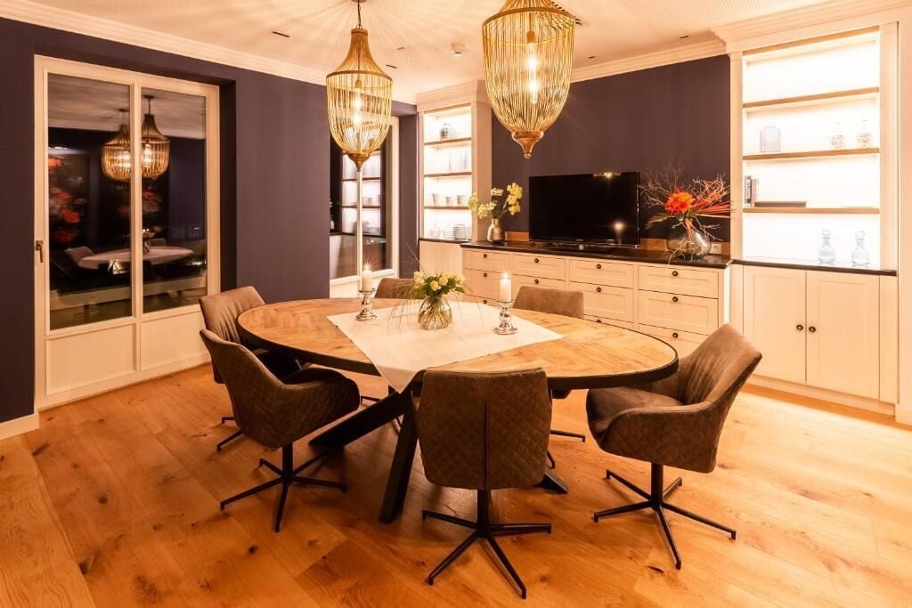 A dining area with a round table, four chairs, a chandelier, and a television in the background.