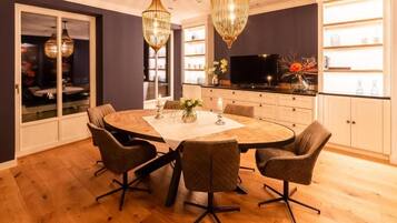 A dining area with a round table, four chairs, a chandelier, and a television in the background.