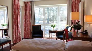 A hotel room with a large bed, two chairs, a small table with a vase, and a view of trees through the window.