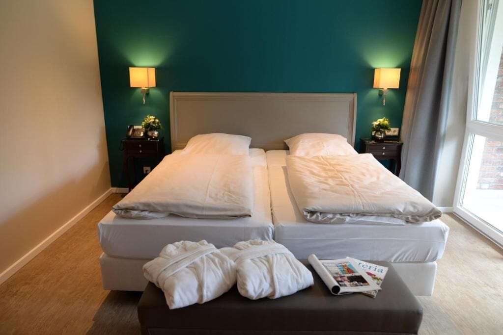 Una cama doble con sábanas blancas, dos mesitas de noche con lámparas, una ventana con cortinas y una revista en el suelo.