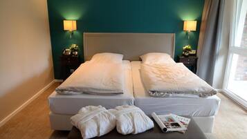 Una cama doble con sábanas blancas, dos mesitas de noche con lámparas, una ventana con cortinas y una revista en el suelo.
