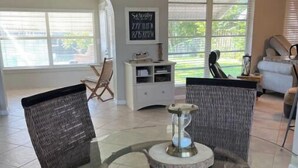 A modern living room with a glass-top dining table surrounded by wicker chairs, a white entertainment unit, and large windows offering a view of the outdoors. - Serenity Canal Cottage 2/2 on Gulf Access Canal!! (North Port)