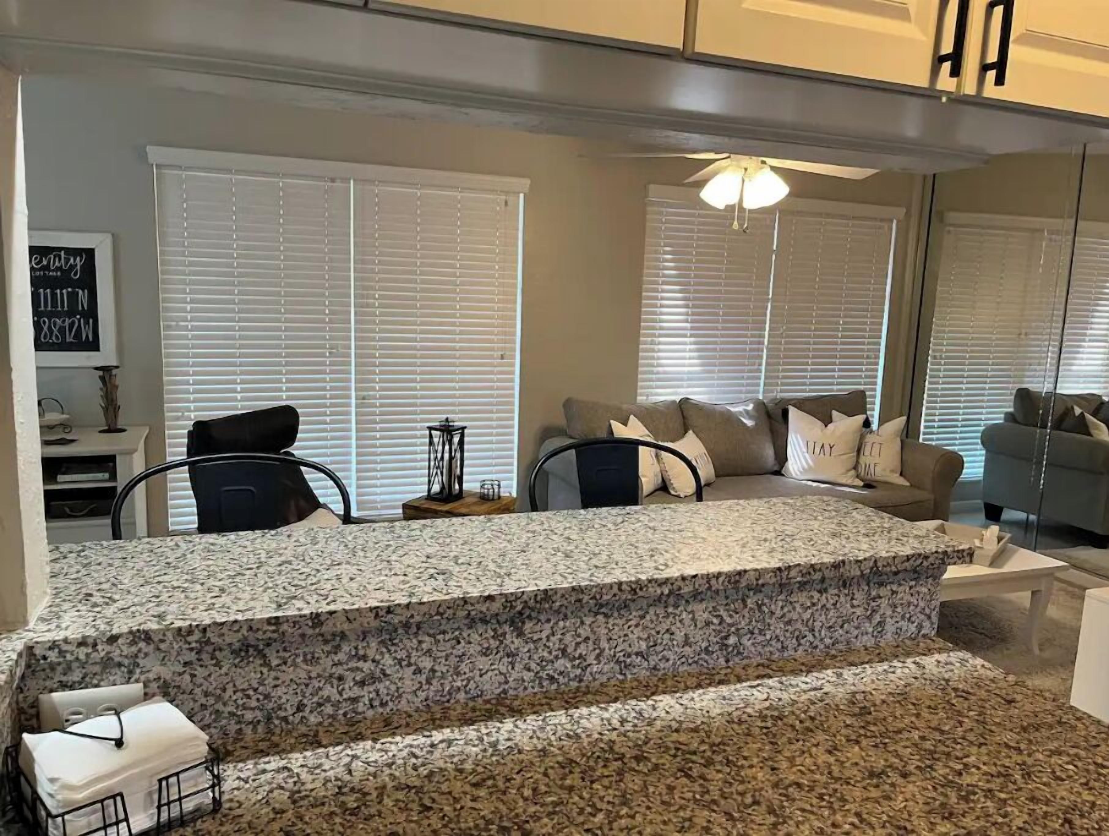 A modern living room with a kitchen island, large windows with blinds, and a ceiling fan.