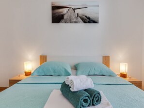 Una cama bien hecha con ropa de cama y almohadas color turquesa, una toalla enrollada y un cuadro con una imagen de un muelle en la pared.