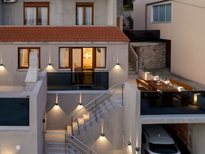 A modern building with a red-tiled roof, a staircase, and a dining area with outdoor lighting. - Rina Apartments - One Bedroom Apartment with Terrace and Sea View (Baric Draga)