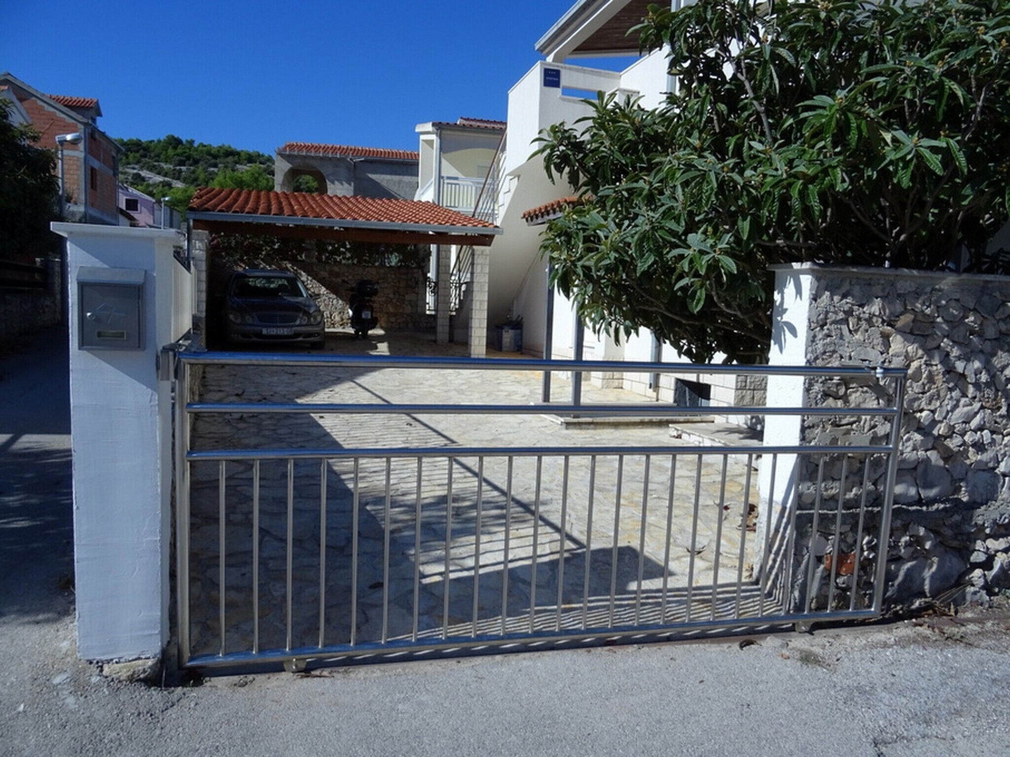Une entrée fermée par un portail donnant accès à une propriété entourée d’un mur en pierre et dotée d’un portail en métal, avec une voiture garée et un bâtiment surmonté d’un toit en tuiles.