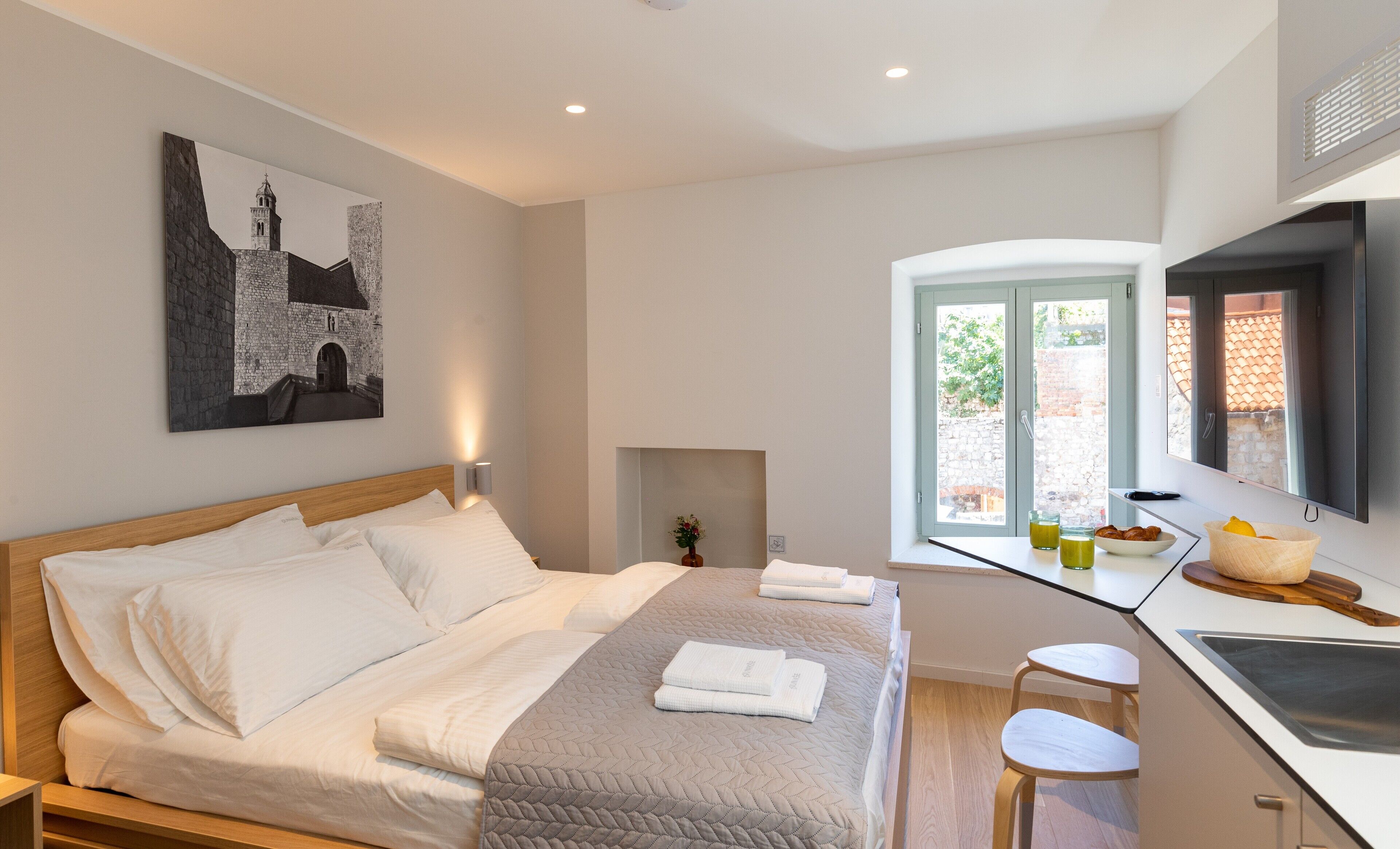 Een moderne hotelkamer met een groot bed, een klein tafeltje met een bloevas, een kitchenette en uitzicht op een tuin via een glazen deur.