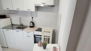 Une petite cuisine avec des meubles blancs, un fourneau et une table avec une chaise. Il y a une plante sur la table.