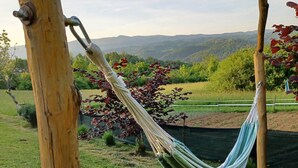 Un hamac est suspendu entre deux poteaux en bois, dans une zone herbeuse, offrant une vue sur des collines et des arbres en arrière-plan.