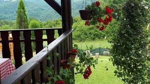 Un balcon en bois, orné d’une nappe à carreaux rouges et blancs, de géraniums en pot, et offrant une vue sur un paysage verdoyant et luxuriant.