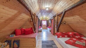 Une pièce mansardée chaleureuse aux murs en bois et aux plafonds inclinés, comprenant deux lits avec une literie rouge, un petit coin salon avec une nappe à carreaux et une fenêtre avec des rideaux.