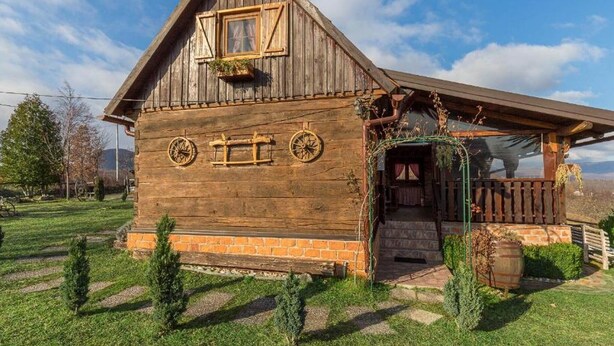 Une maison rustique en bois, dotée d’un toit en pente, d’un petit porche avec une balustrade en bois et d’une fenêtre agrémentée d’un pot de fleurs suspendu.