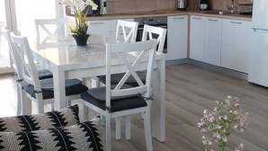 Eine moderne Küche mit weißen Schränken, ein Essbereich mit weißen Stühlen sowie ein Wohnzimmer mit einer Couch und einem Couchtisch.
