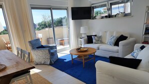 Una sala de estar moderna con un sofá blanco, un sillón estampado y una mesa de centro redonda. Un ventanal ofrece vistas al exterior.