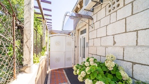 Un sendero estrecho que conduce a una puerta blanca con una reja metálica decorativa, rodeado de follaje y un felpudo de ladrillo.