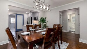 Uma sala de jantar com uma mesa de madeira posta para seis pessoas, rodeada por cadeiras, um lustre moderno e uma planta em vaso.