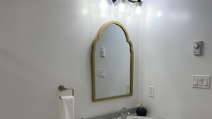 Ein Badezimmer mit einer weißen Toilette, einem Waschbecken mit spiegel und goldener Rahmung darüber sowie einer Wandleuchte.
