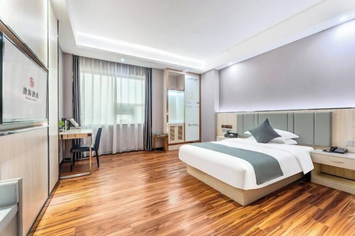 A hotel room with a wooden floor, a bed with a grey headboard, a desk with a chair, and a large window with curtains.
