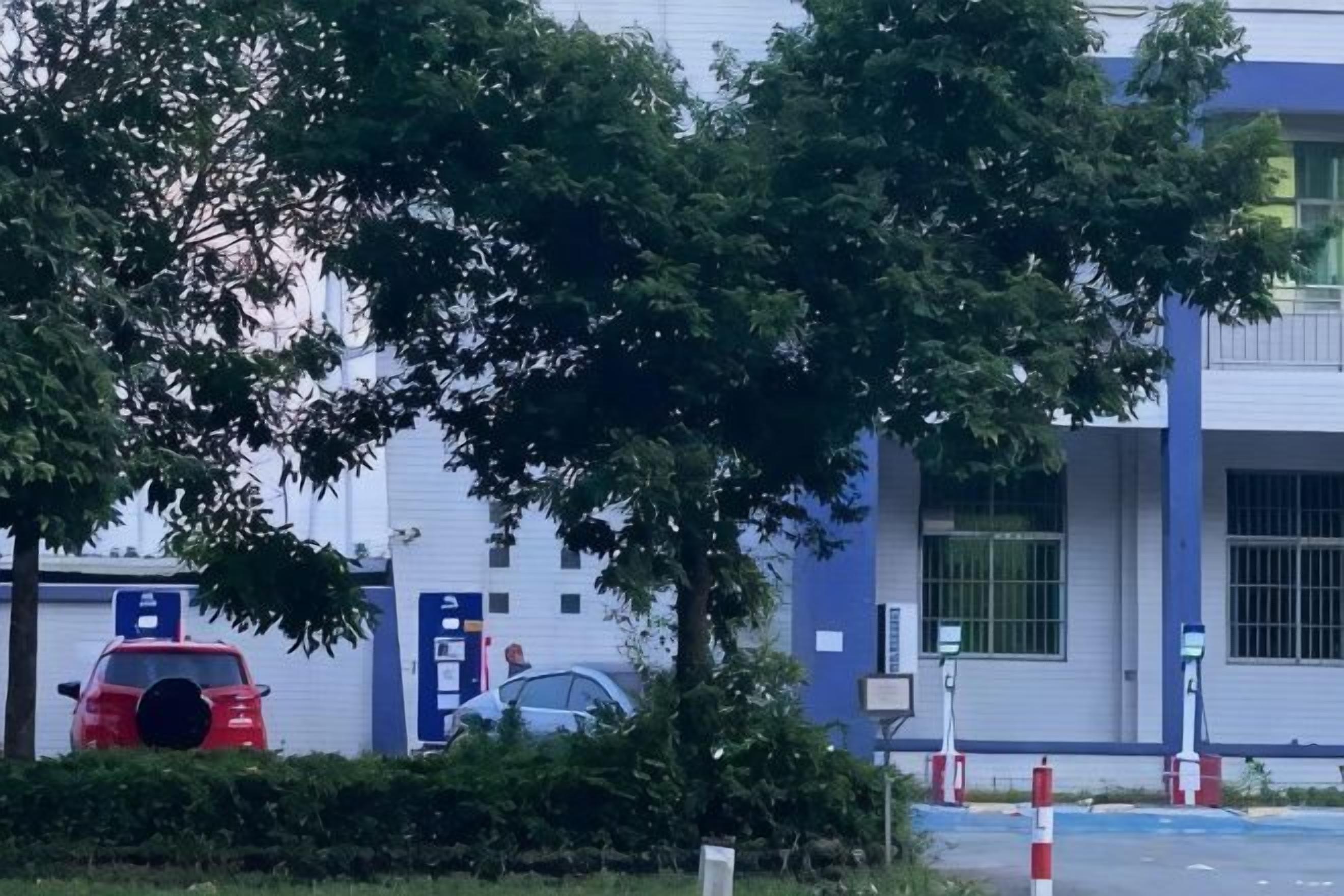 A parking area with cars, a building with blue and white exterior, and trees.