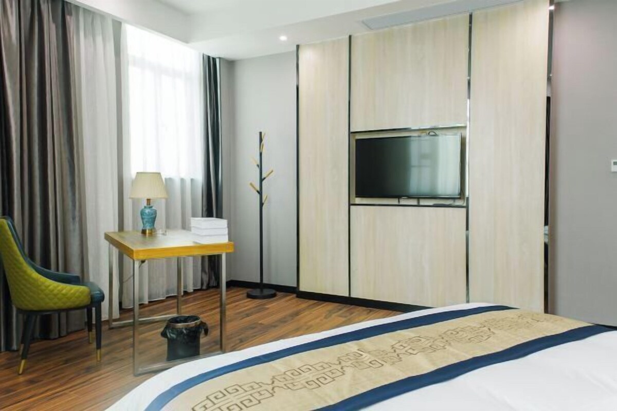 A hotel room with a bed, a desk with a lamp, a chair, a TV on the wall, and a trash bin.