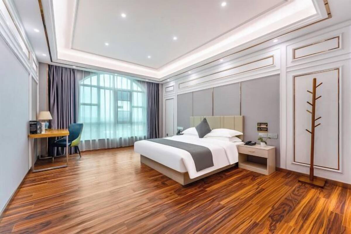 A modern hotel room with a wooden floor, a large bed with a grey and white duvet, a bedside table with a lamp, and a view of the city through large windows.