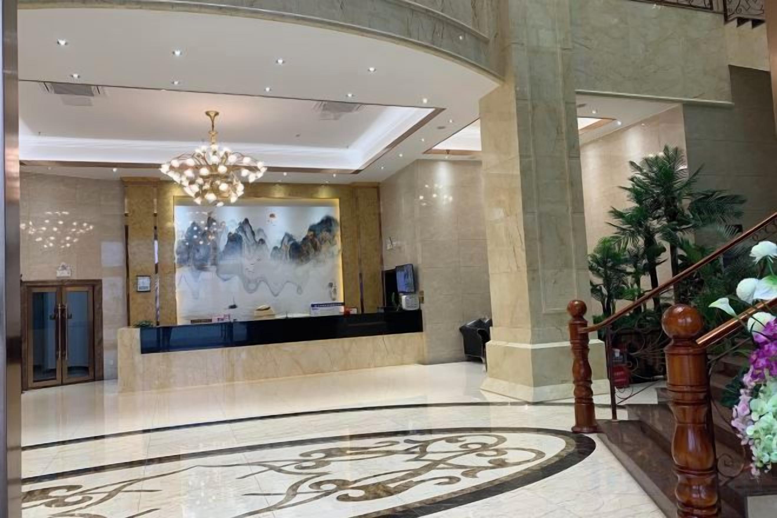 A hotel lobby with a reception desk, a chandelier, a large wall painting, and a staircase.
