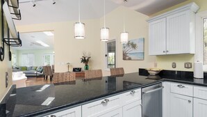 A modern kitchen with white cabinets, a black countertop, and pendant lights. There is a dining area with wicker furniture and a painting of a beach scene. - 3 BR/2BA Home! Screen Porch! Amenity access included! (Seabrook Island)