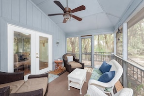 A covered porch with wicker furniture, a ceiling fan, and large windows overlooking a wooded area.