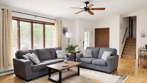 Ein Wohnzimmer mit zwei grauen Sofas, einem Holztisch, einem gemusterten Teppich, einem Deckenventilator und einer Treppe.