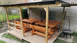 Een houten eethoek buiten met banken en tafels, een pergola erboven en een barbecuehoek.