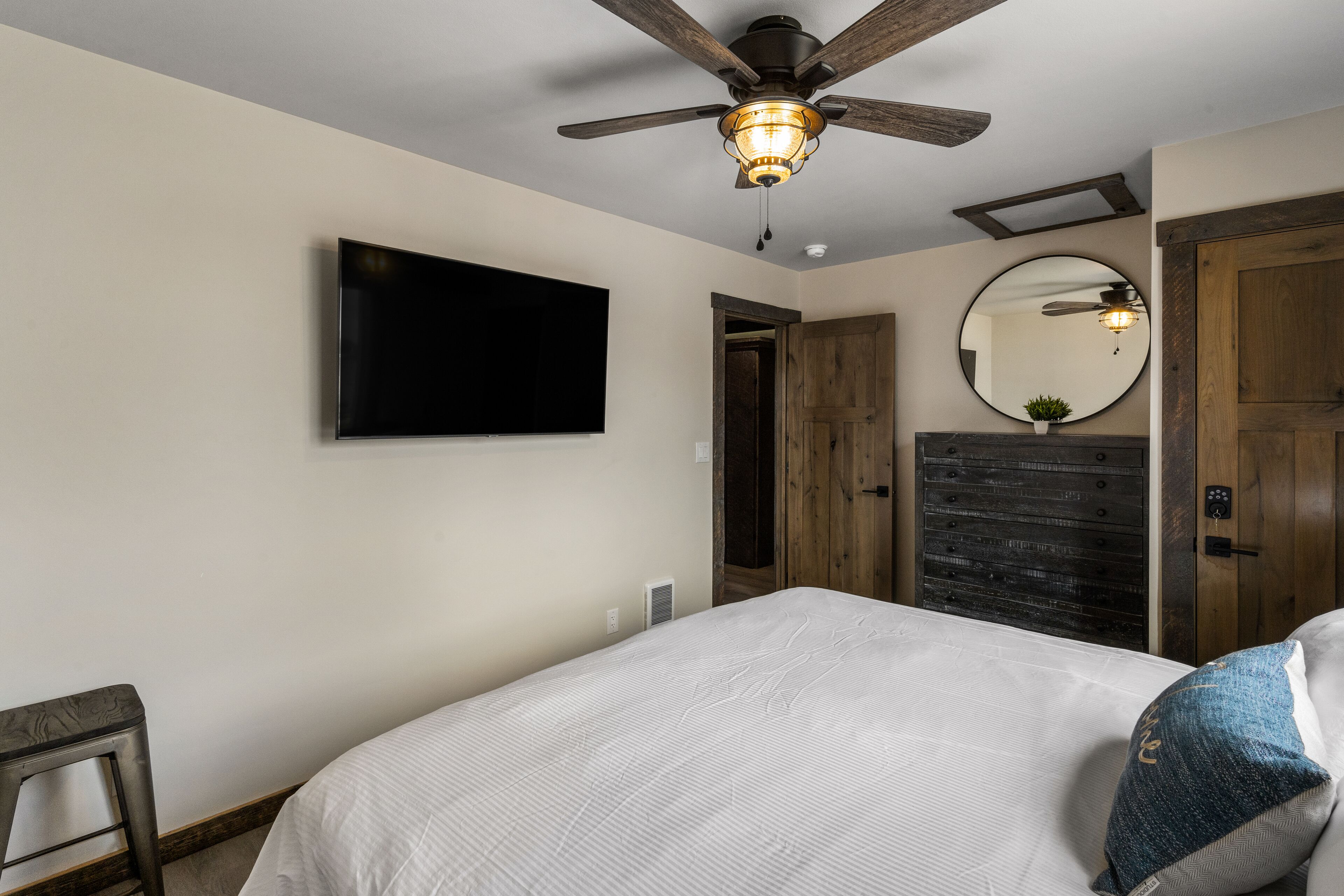 A bedroom with a bed, a ceiling fan, a mounted TV, a wooden dresser, and a mirror.