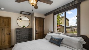 A bedroom with a large bed, wooden wardrobe, a ceiling fan, a round mirror, and a window with a view of a building and trees. - Little Tuckaway (West Yellowstone)