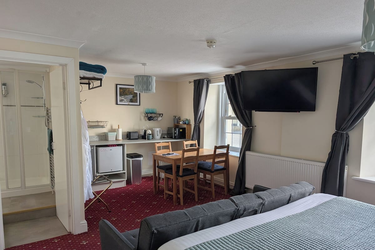 A hotel room with a bed, a desk with a chair, a kitchenette, a TV, and a bathroom with a shower.