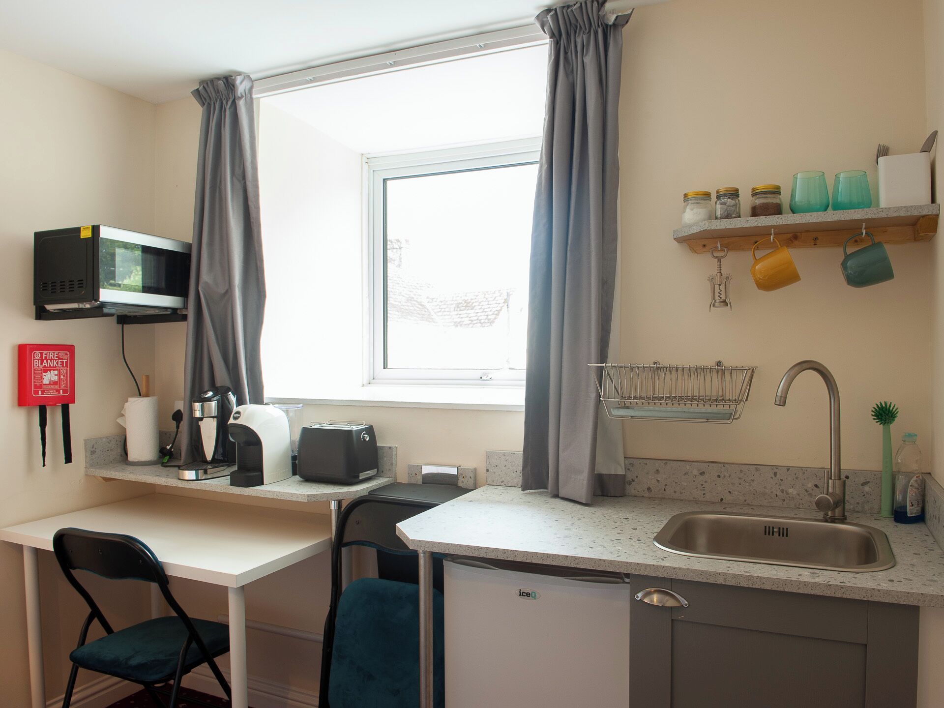 A compact kitchen with a microwave, toaster, and sink. A fire blanket is mounted on the wall, and a window with curtains is visible.