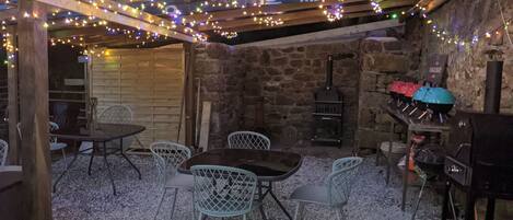 Un espacio comedor al aire libre de estilo rústico con pared de piedra, vigas de madera y luces colgantes.