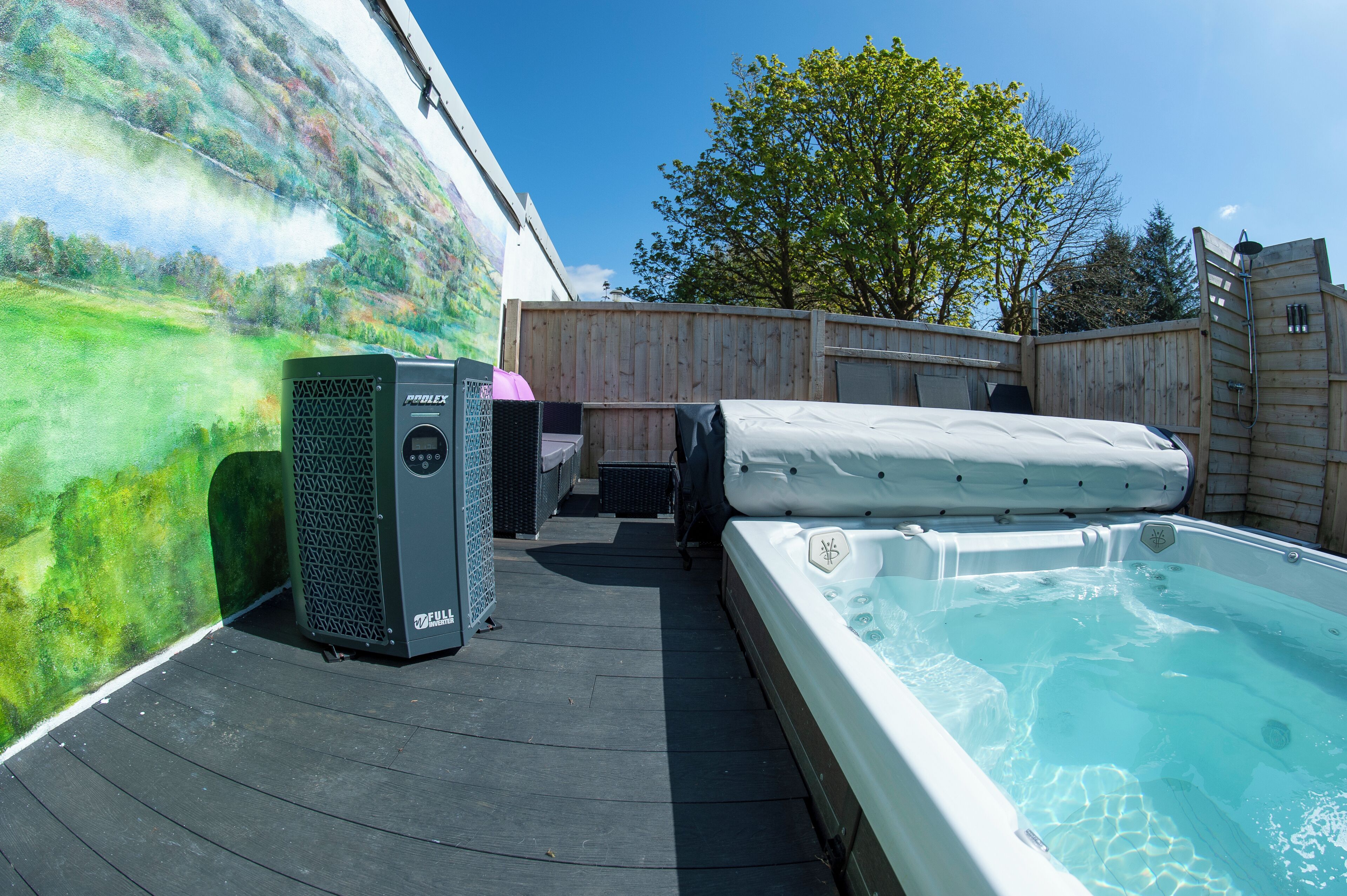 A hot tub and a spa heater on a wooden deck with a mural wall and a wooden fence in the background.