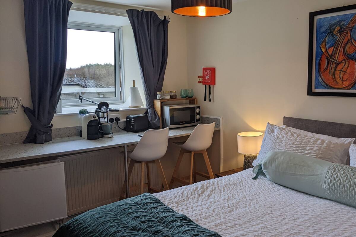A hotel room with a bed, a desk with a computer, a chair, a window with curtains, and a painting on the wall.